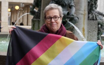Foto von Sofie Langmeier, sozialpolitische Sprecherin Die Grünen/Rosa Liste/Volt, mit der Disability Pride Fahne vor dem Fischbrunnen