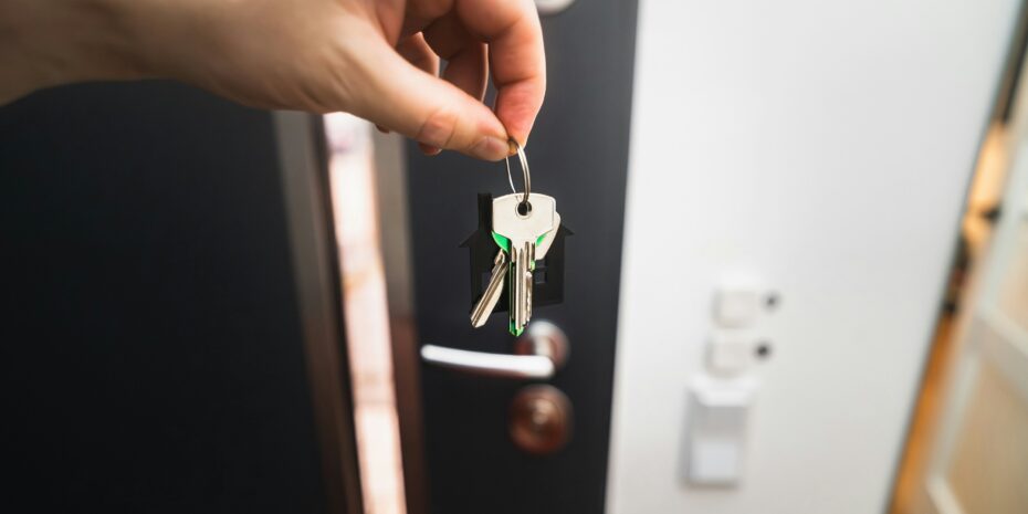 Aufnahme von einer Hand, die Wohnungsschlüssel hält; im Hintergrund ist eine Wohnungseingangstür zu sehen